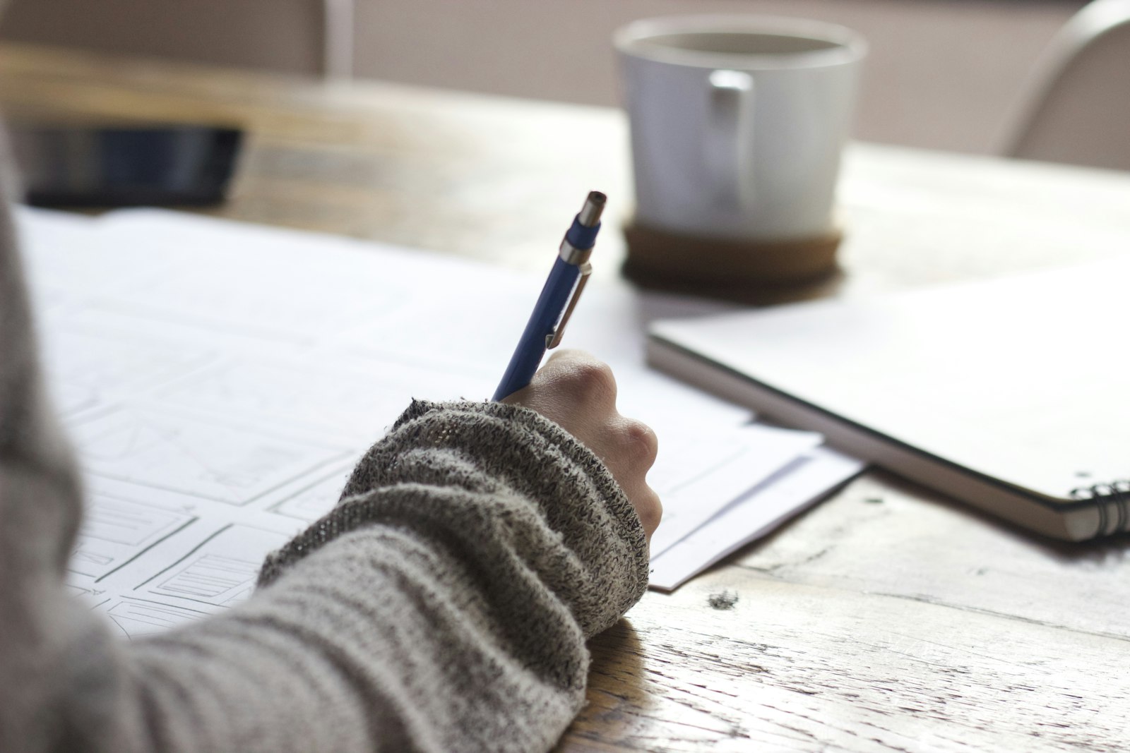 A hand writing on printed papers at a café table beside a coffee cup